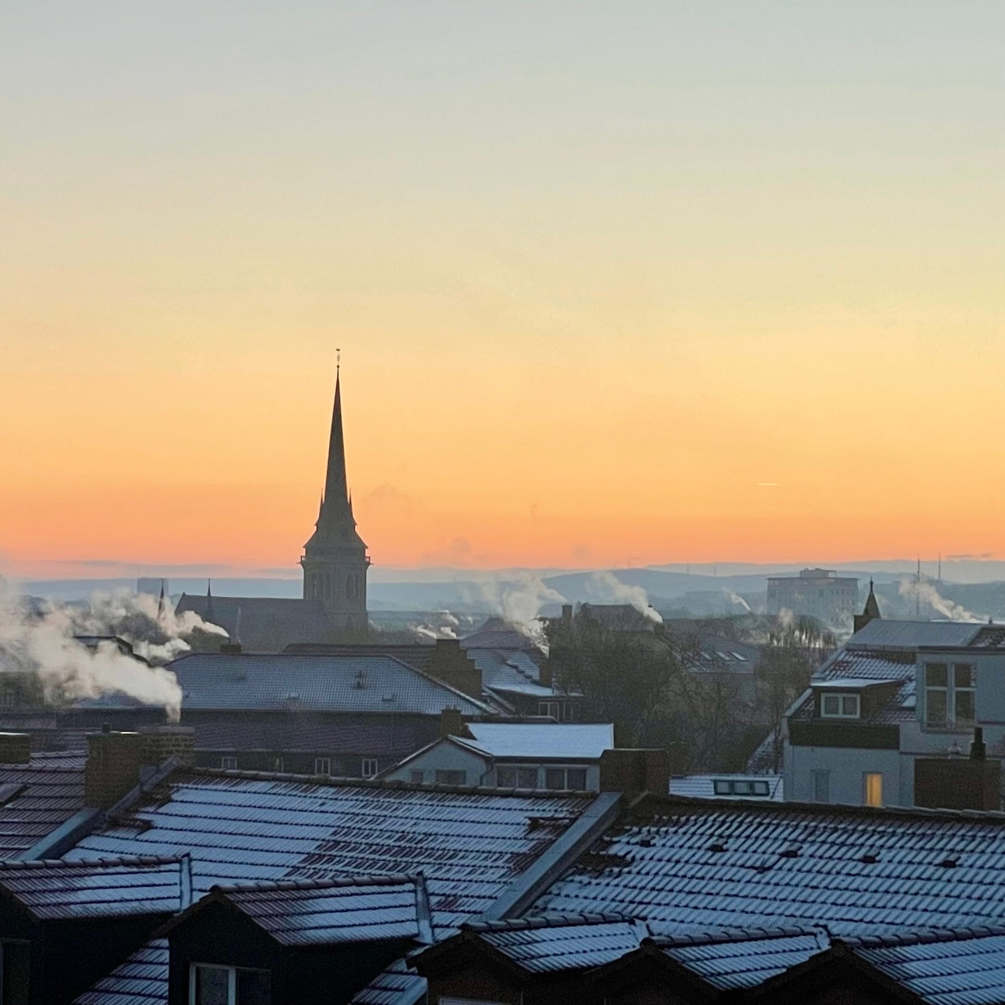 Der Himmel leuchtet rot und eine Kirchturmspitze ist da auch. Die Dächer sind Winter und Schnee bedeckt, überall rauchen die Schornsteine.